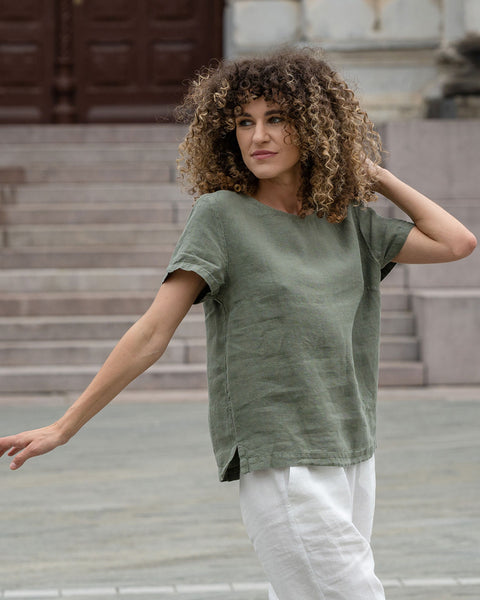 Hermosa mujer con cabello rizado en la ciudad, posando con una blusa de lino verde piedra Emma y pantalones de lino blanco puro Dakota.