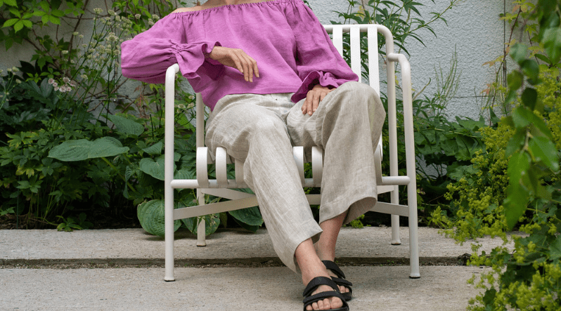 a woman, sitting outside in a chair in a garden, wearing a long sleeve pink ruffle blouse, and natural color linen pants.