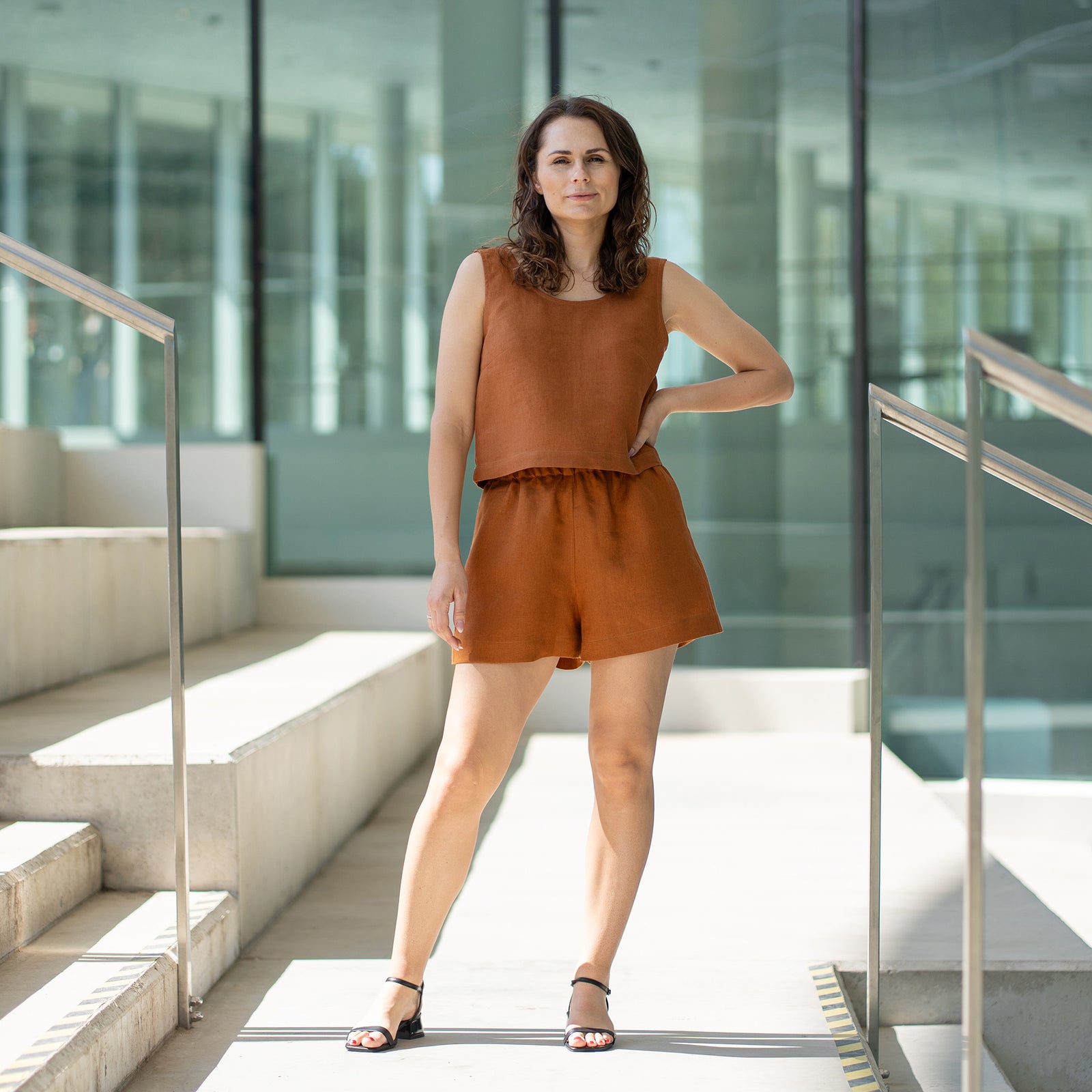 Una mujer de cabello castaño, vestida con un conjunto a juego de blusa sin mangas y pantalones cortos color almendra, mira por encima del hombro mientras está de pie en unas escaleras exteriores de concreto con barandillas de metal.