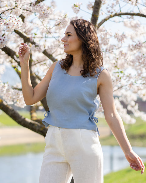 Una mujer con cabello largo, ondulado y castaño lleva una blusa sin mangas azul grisáceo con lazos laterales y pantalones blancos de lino, mirando hacia la derecha. Está de pie al aire libre bajo un cerezo en flor, con un cuerpo de agua tranquilo y un paisaje verde al fondo.