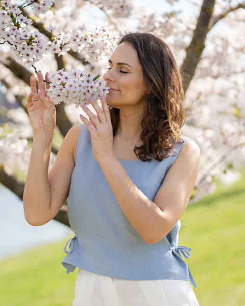 Una mujer de cabello castaño y ondulado, vestida con una blusa sin mangas azul grisáceo, está de pie con los ojos cerrados, acercando suavemente una rama de flores blancas de cerezo a su nariz como si quisiera olerlas. Al fondo, desenfocado, se ve un cuerpo de agua azul claro y hierba verde.
