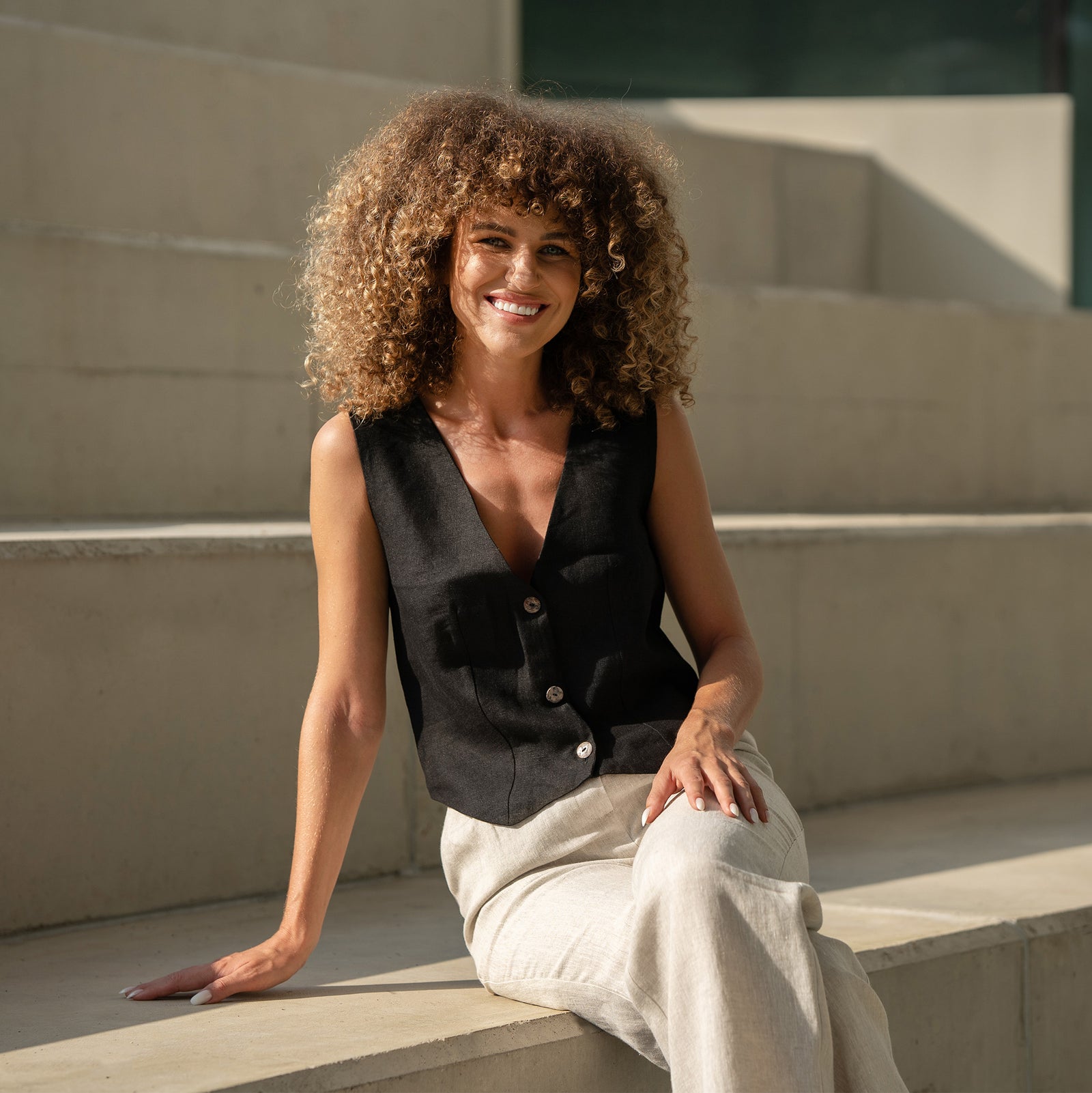 Chaleco de lino Ava en negro combinado con pantalones de lino color natural. Mujer caminando y sonriendo