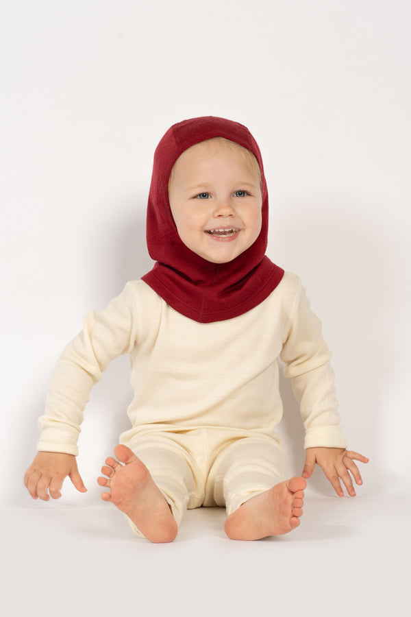 Una niña pequeña sentada en el suelo, vistiendo la braga para la cabeza de merino 160gsm para niños de menique en color cereza real, imagen con fondo de estudio.