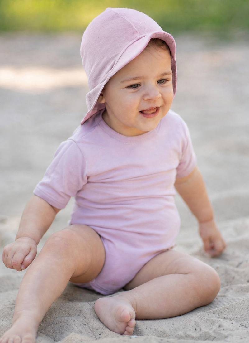 A smiling baby sitting barefoot in the sand outdoors, looking happily off to the side. The baby is wearing a light pink short-sleeve bodysuit and a matching dusty pink sun cap with a front brim and protective neck flap.