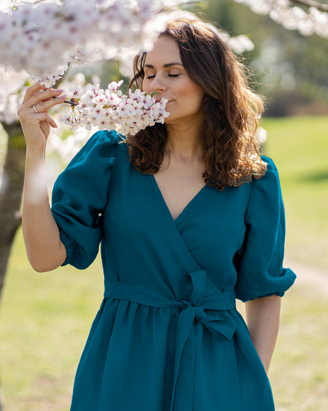 Una persona de piel clara con cabello castaño ondulado tiene los ojos cerrados y parece estar oliendo un racimo de flores blancas que sostiene en su mano derecha. Lleva un vestido cruzado de lino Menique con mangas cortas abullonadas y cintura atada, y su mano izquierda está en un bolsillo. Un árbol en flor y un fondo verde desenfocado sugieren un entorno al aire libre.