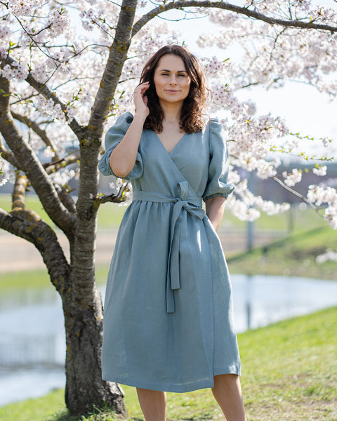Una mujer de cabello castaño, vestida con un vestido cruzado de lino verde menta con mangas abullonadas hasta el codo, está de pie al aire libre junto a un árbol con flores blancas.