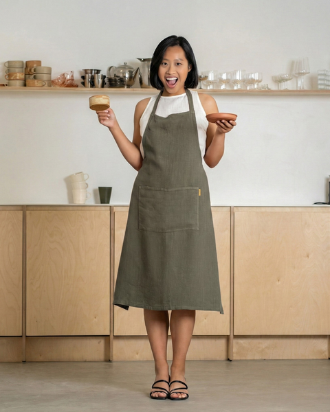 Mujer con un delantal de lino verde piedra sosteniendo una taza y un plato, sonriendo en un interior de cocina minimalista.