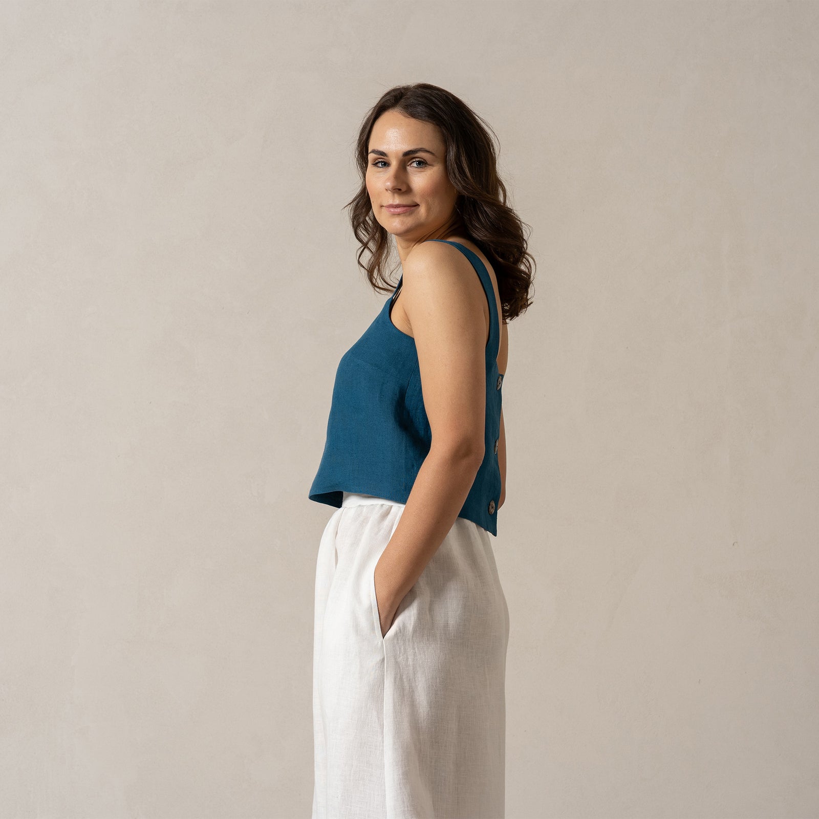 Una mujer de piel de tono medio y cabello oscuro mira directamente a la cámara con una expresión neutral. Lleva una blusa sin mangas de lino color verde azulado con escote cuadrado y una falda de lino blanco puro de menique, con las manos en los bolsillos de la falda. El fondo es de un color beige claro y liso.