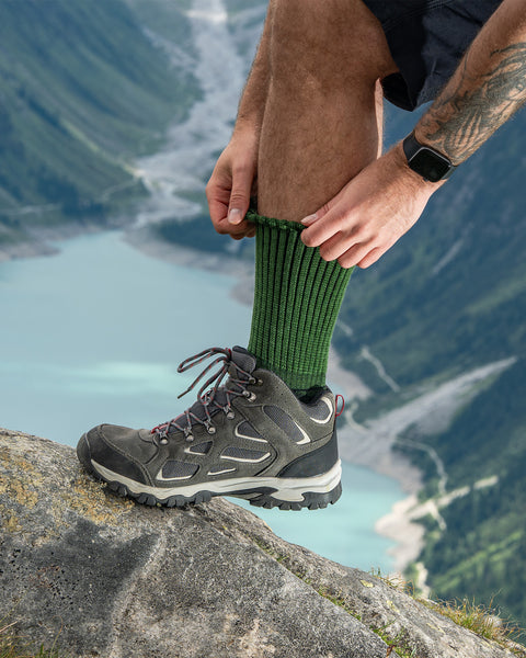 Hombre ajustándose calcetines verdes de merino acanalados para senderismo mientras lleva botas de montaña, con un paisaje de lago alpino pintoresco al fondo.