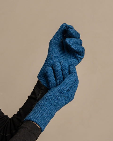 Una toma en primer plano de las manos de una persona que lleva guantes de punto acanalado de color azul vibrante de la marca menique. La mano izquierda está cerrada en un puño, mientras que la mano derecha parece sujetar la zona de la muñeca del guante izquierdo. La persona lleva una camisa oscura de manga larga debajo. El fondo es de un gris neutro y uniforme.