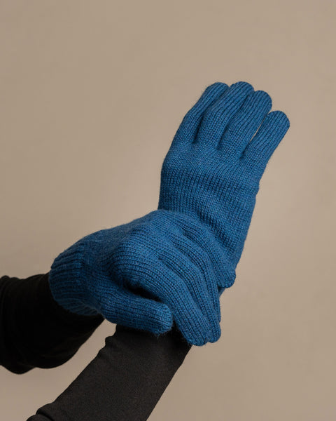 Una toma en primer plano de las manos de una persona que lleva guantes de punto acanalado de color azul vibrante de la marca menique. La mano derecha sostiene suavemente la parte inferior de la mano izquierda, que está ligeramente en forma de copa. La persona lleva una camisa oscura de manga larga debajo. El fondo es de un gris neutro y uniforme.