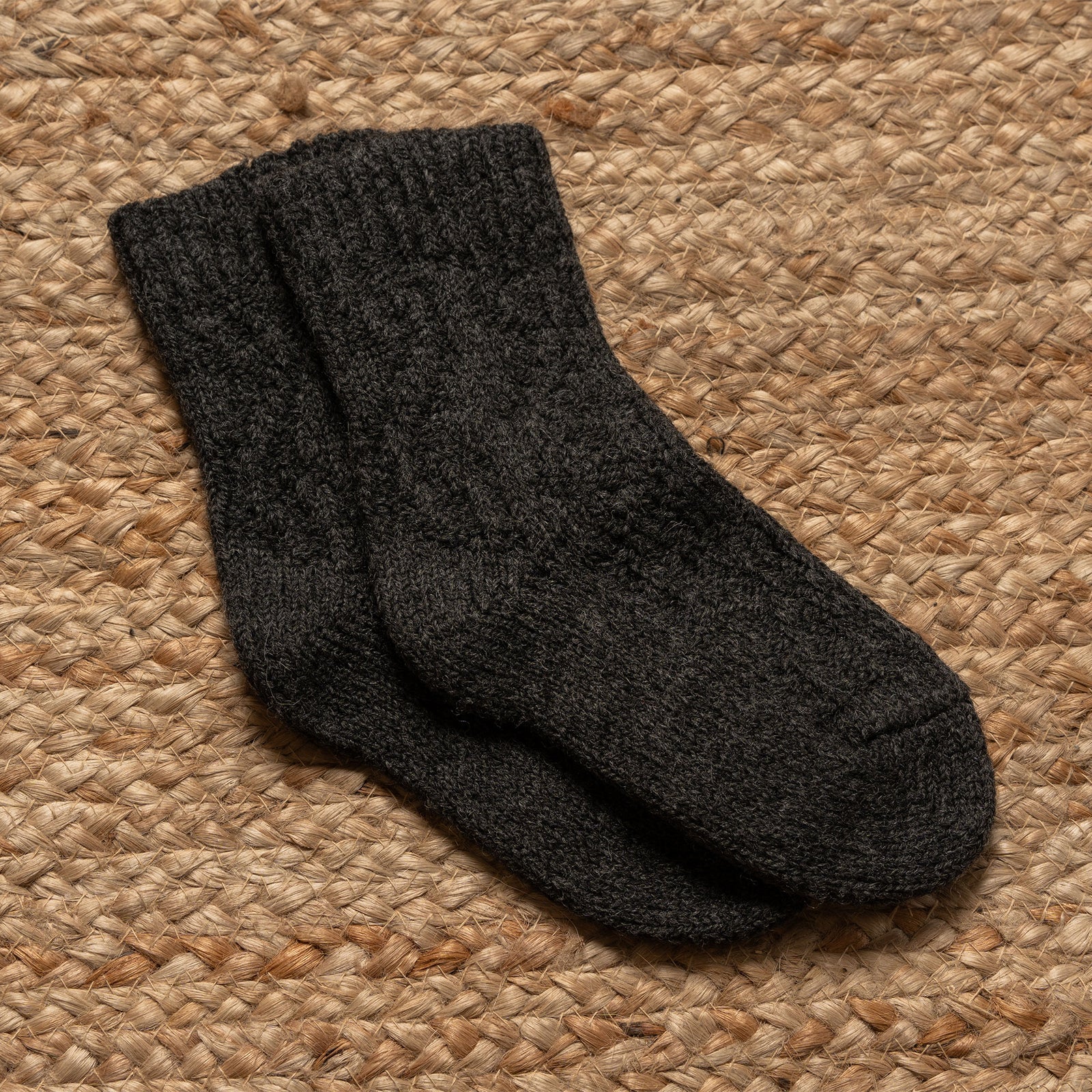 Una toma desde un ángulo bajo muestra las piernas y los pies de una persona, probablemente usando calcetines de lana tejidos de la marca menique en color blanco roto, de longitud hasta el tobillo, con un puño acanalado visible y un patrón texturizado de trenzas en el pie. La persona está de pie sobre una estera tejida de color marrón claro, con un fondo neutro y liso.
