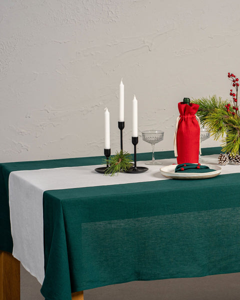 El mantel de lino menique en color verde oscuro, decorando una mesa de Navidad. Hay elementos naturales, como piñas y ramas de pino sobre la superficie de la mesa. También hay una botella de vino en una bolsa de almacenamiento y velas.