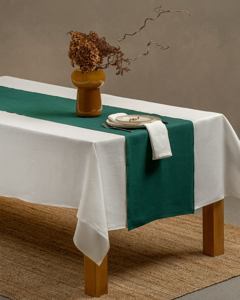 El mantel de lino menique en color blanco puro y el camino de mesa en color verde oscuro, cayendo sobre una mesa de madera, con un jarrón de cristal y una taza de té con un plato encima.