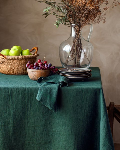 Mantel de lino verde oscuro decorado con frutas rústicas y cesta tejida sobre una mesa de comedor.