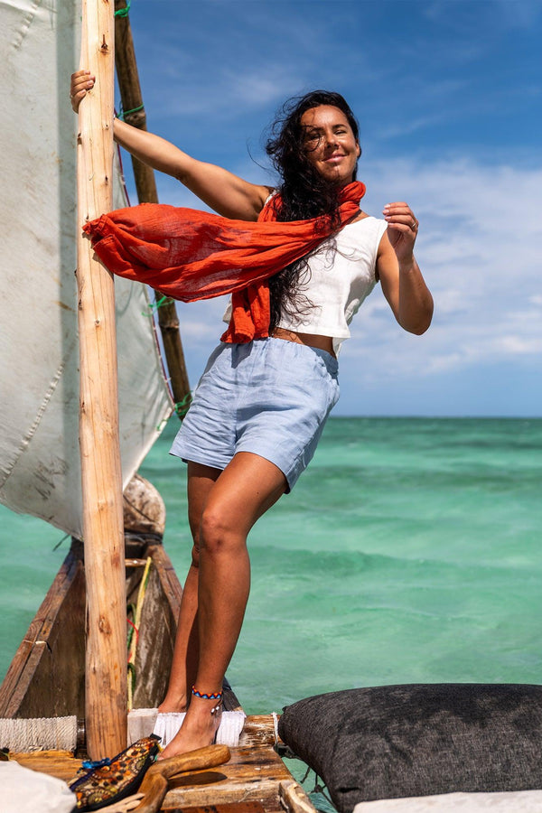 Una mujer está de pie en un bote rústico de madera con una vela blanca contra un cielo azul brillante y agua turquesa. Lleva una blusa blanca sin mangas, pantalones cortos azul claro y un pañuelo naranja. El bote tiene un cojín blanco y un cojín gris oscuro.