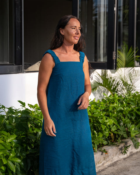 Una mujer de cabello largo y oscuro está al aire libre, vistiendo un vestido de lino para el sol con escote cuadrado y tirantes gruesos. Ella mira hacia un lado, rodeada de frondosa vegetación verde. Al fondo se ve un edificio blanco con ventanas de marco oscuro y más vegetación.