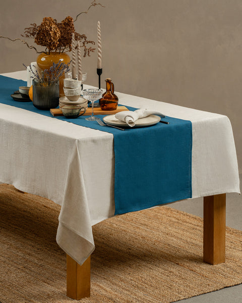 Una vista de una mesa de madera cubierta con un mantel blanco, con un camino de mesa azul cobalto encima. Hay jarrones, tazas y otras cerámicas sobre ella. La mesa está colocada sobre una alfombra, con un fondo de estudio.