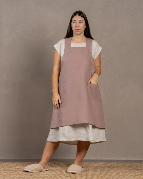 Una mujer que lleva un delantal de lino color rosa desvaído sobre un vestido de lino menique color natural, de pie frente a un fondo texturizado.