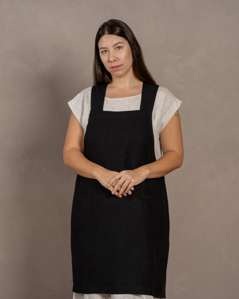 Una joven de cabello oscuro de pie frente a un fondo de estudio, con las manos juntas delante. Lleva un vestido de lino con un delantal japonés cruzado de lino en color negro puro, talla L/XL, encima.