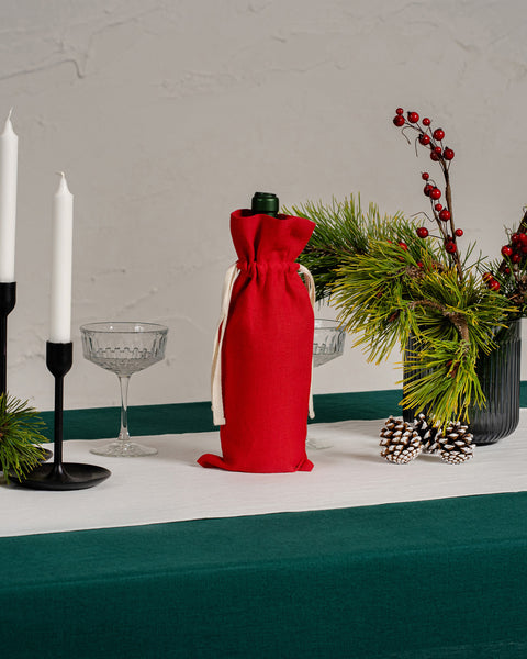 Decoración elegante para la mesa de Navidad con un mantel de lino verde oscuro, camino de mesa de lino blanco, bolsa para vino de lino rojo, cristalería, candelabros negros, ramas de pino, bayas rojas y piñas.