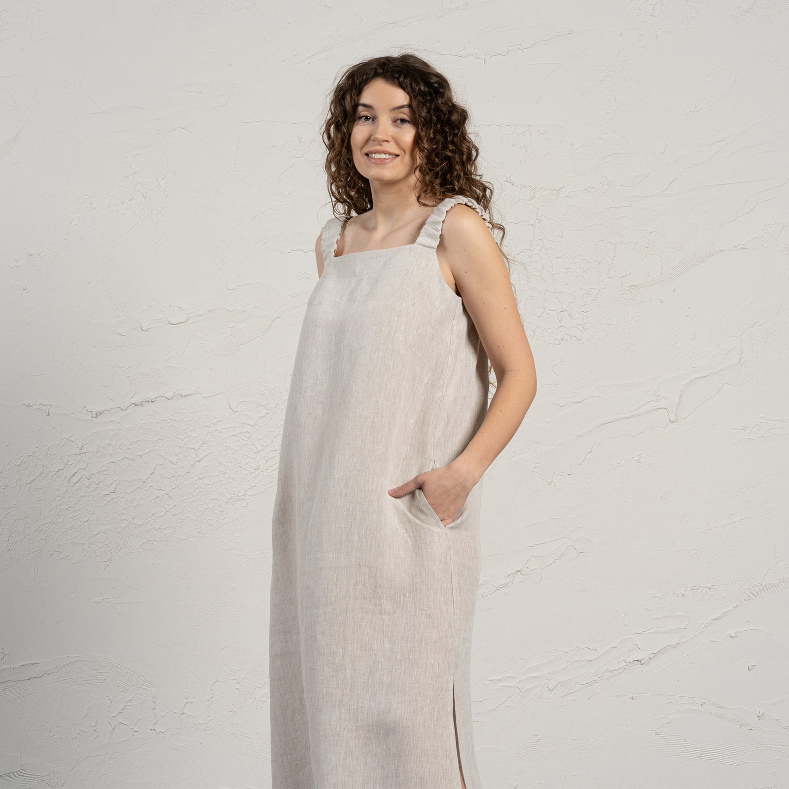 Una mujer de piel clara con cabello largo, rizado y oscuro está de pie mirando a la cámara con una sonrisa amable. Lleva un vestido largo sin mangas de Menique con escote cuadrado y aberturas laterales, y sandalias negras. Tiene las manos en los bolsillos del vestido y está sobre un suelo gris claro frente a una pared texturizada de color blanco roto.