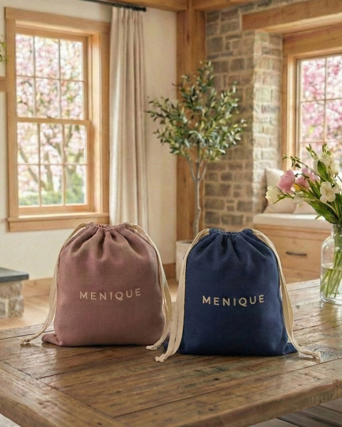 Dos bolsas de regalo con cordón de la marca Menique en tonos rosa empolvado y azul marino, colocadas sobre una mesa de madera en una sala de estar cálida y bañada por el sol, mostrando un embalaje de primera calidad para regalos de lana merina.