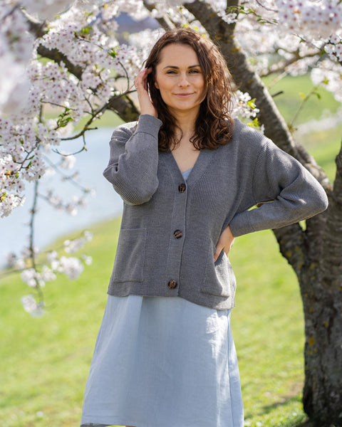 Una mujer de cabello castaño, que lleva un cárdigan gris de punto sobre un vestido azul claro, está de pie al aire libre junto a un árbol con flores blancas.