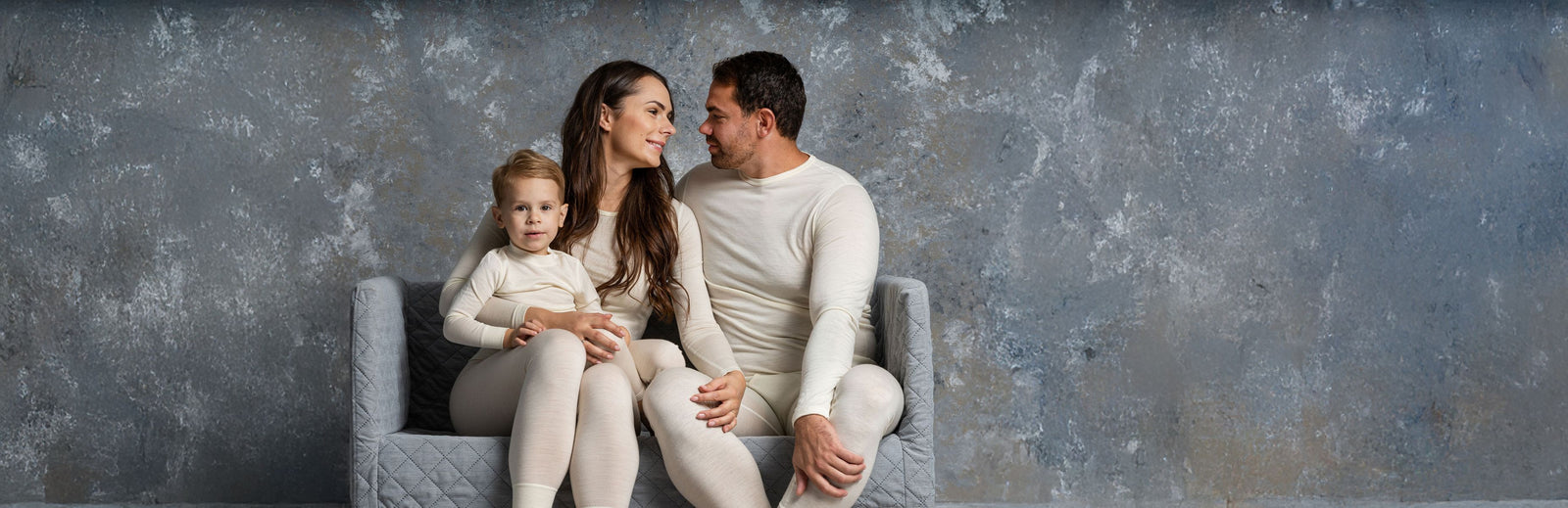 Familia de tres personas sentadas juntas en una silla gris frente a una pared gris con textura.