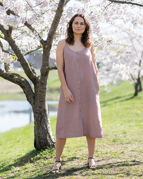 Una mujer con cabello largo, oscuro y ondulado, vestida con un vestido midi sin mangas, con botones al frente y de color rosa desvaído, y sandalias negras abiertas en los dedos, está de pie al aire libre frente a un árbol con flores blancas. Tiene las manos relajadas a los costados. Mira directamente al espectador con una expresión neutral o una leve sonrisa. El fondo es un entorno soleado al aire libre con césped verde y un cuerpo de agua, posiblemente un estanque o lago, y más árboles en flor.