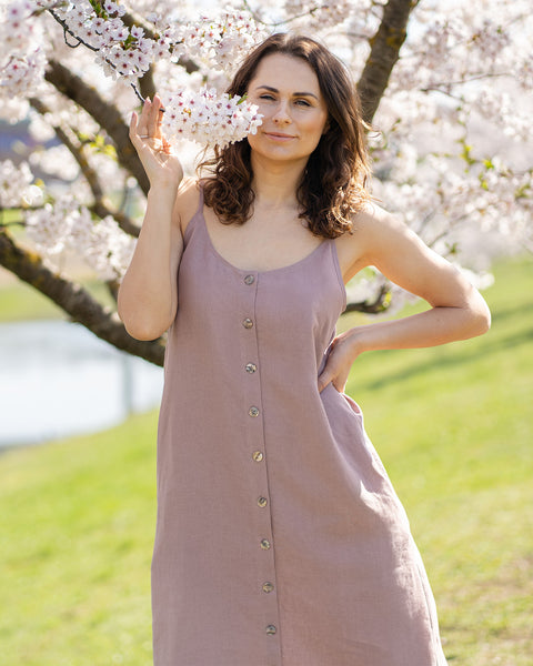 Una mujer con cabello largo, oscuro y ondulado, vestida con un vestido midi sin mangas, con botones al frente y de color rosa desvaído, está de pie al aire libre frente a un árbol con flores blancas. Sostiene una ramita de las flores cerca de su nariz con la mano derecha. Su mano izquierda está en la cadera. Mira directamente al espectador con una leve sonrisa. El fondo es un entorno soleado al aire libre con césped verde y más árboles en flor.