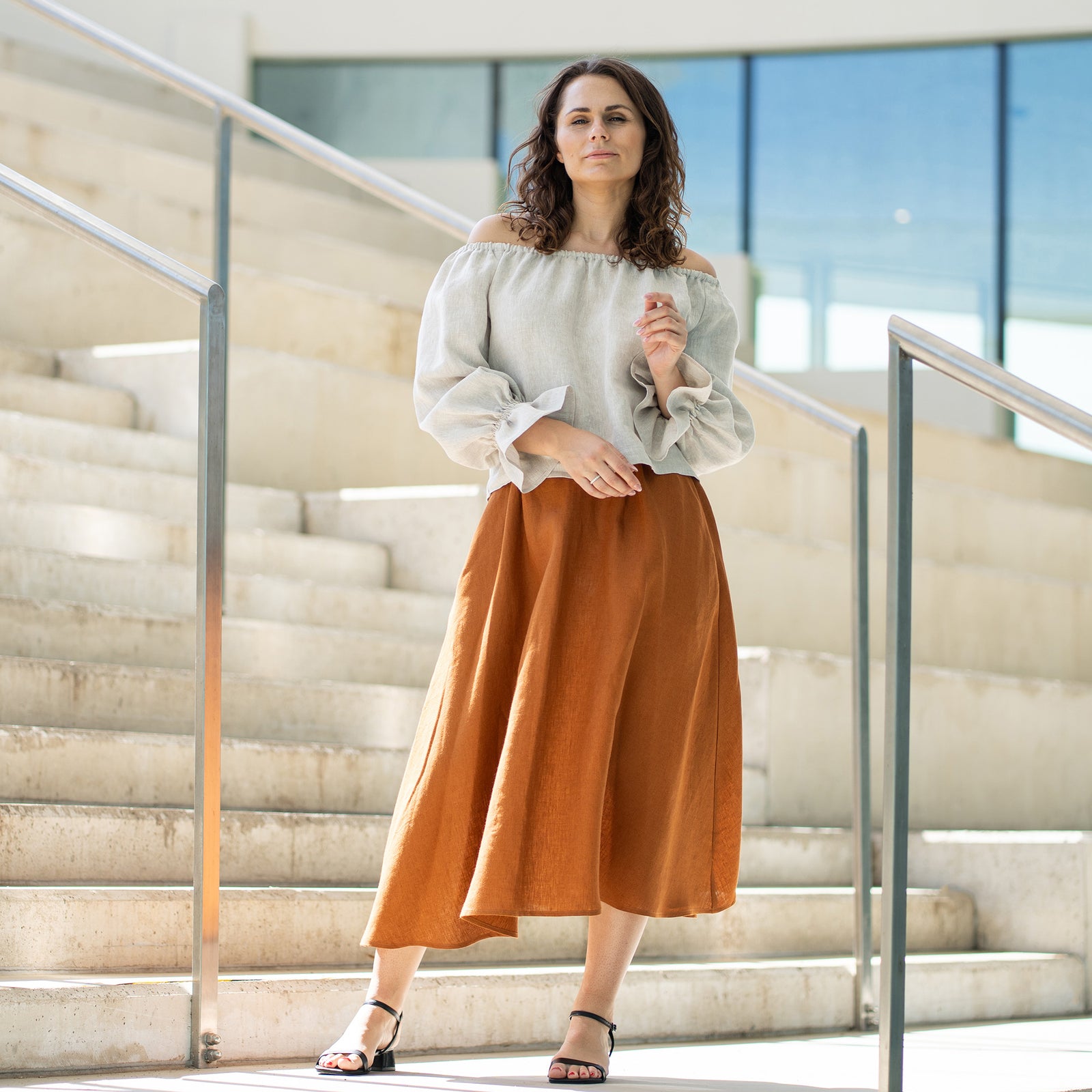 Una mujer de cabello castaño, vestida con una blusa beige claro con los hombros descubiertos y mangas abullonadas, y una falda larga color óxido, posa sobre unas escaleras de cemento.