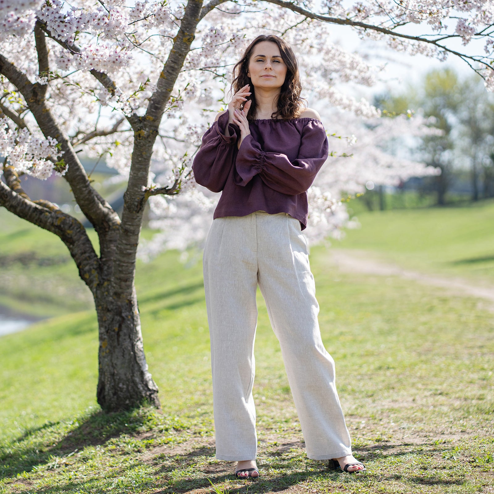 Se muestra a una mujer de piel clara y cabello oscuro desde la cintura hacia arriba, sonriendo directamente al espectador. Lleva una blusa de cuello redondo en tono púrpura sombra. Su mano izquierda, adornada con un anillo en el dedo anular, está levantada de manera casual hacia su pecho, con el pulgar tocando su escote.