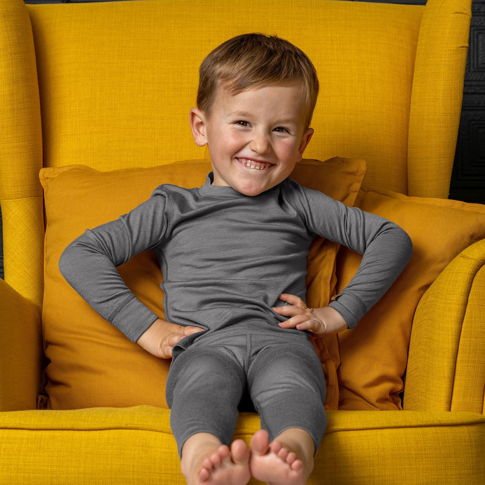 Niño sonriente con conjunto de capa base de lana merina gris, sentado con las piernas cruzadas en una silla y los brazos levantados.