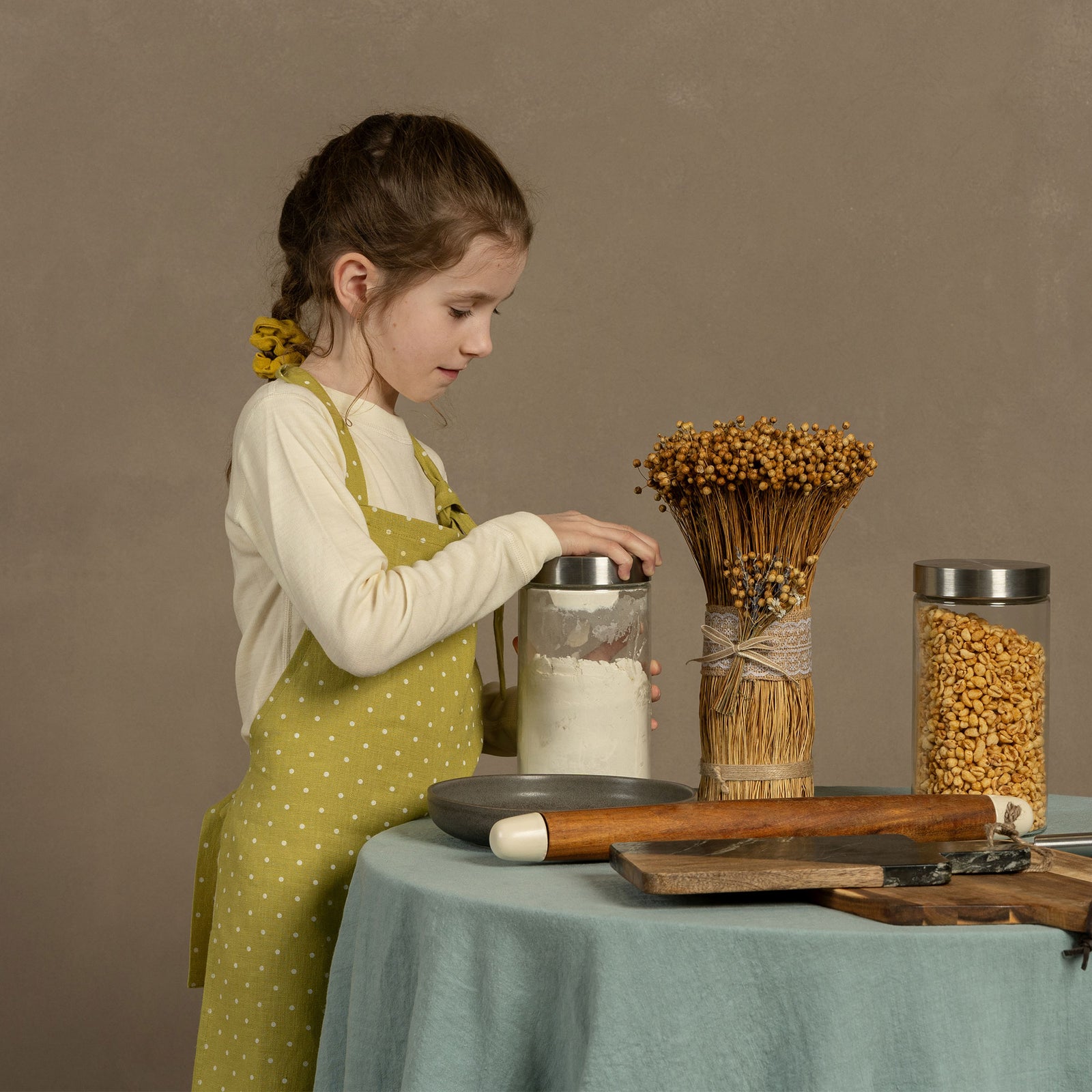 Un niño con un delantal de lino con puntos en forma de hoja, de pie junto a una mesa participando en manualidades o repostería, con ingredientes y utensilios de cocina sobre la mesa.