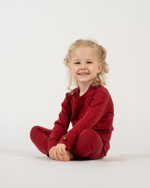 A young child with light curly hair, pulled into pigtails, is sitting on the floor with their legs crossed and a bright smile on their face. They are wearing a long-sleeved red top and matching red pants. The background is white.