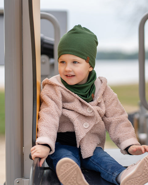 Child sitting on a playground slide, dressed in a dark green Merino wool beanie and neck gaiter with a beige coat and jeans.