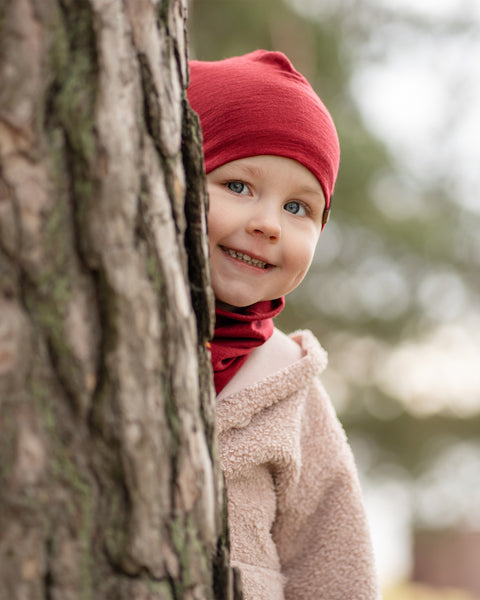 Smiling child peeking from behind a tree, wearing a red Merino wool beanie and matching neck gaiter, outdoor forest background.