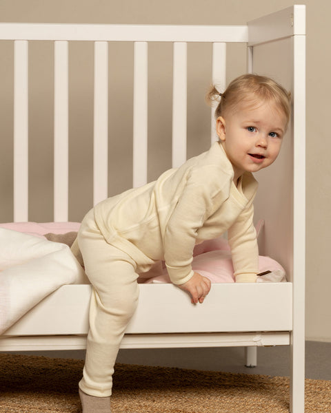 A toddler with blonde hair tied in two small pigtails is standing with one leg inside a white crib and the other leg outside, appearing to climb out. The child is wearing menique long-sleeved top and matching pants, and is looking towards the viewer with a slight smile. Inside the crib, there's a pink blanket and pillow.