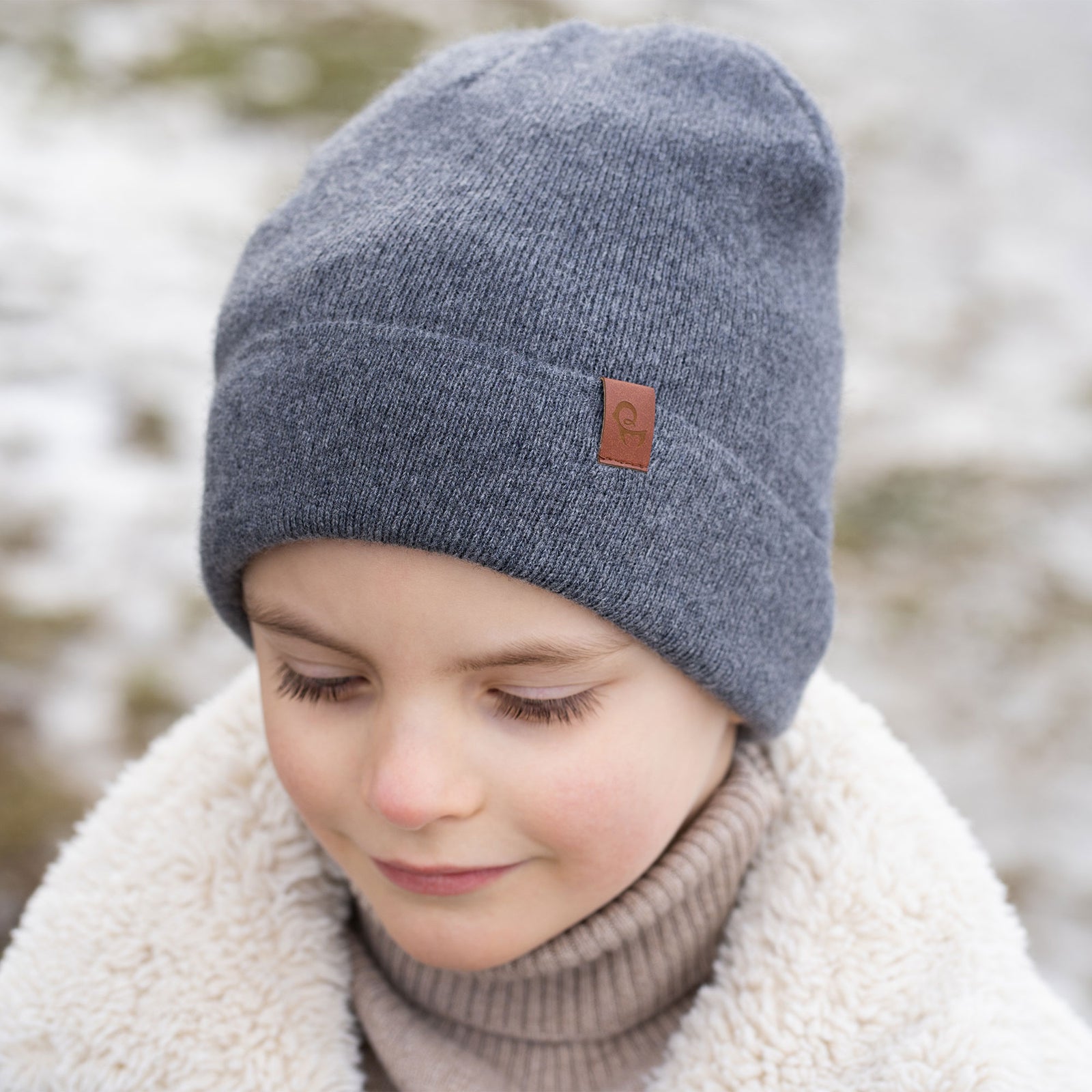 Un niño pequeño con cabello corto y castaño y una amplia sonrisa, está vestido con ropa de invierno. Lleva un gorro beige, un suéter de cuello alto beige y una chaqueta sherpa color crema, con guantes tejidos beige. Detrás de él se ve un fondo nevado y desenfocado con lo que parece ser un cuerpo de agua.