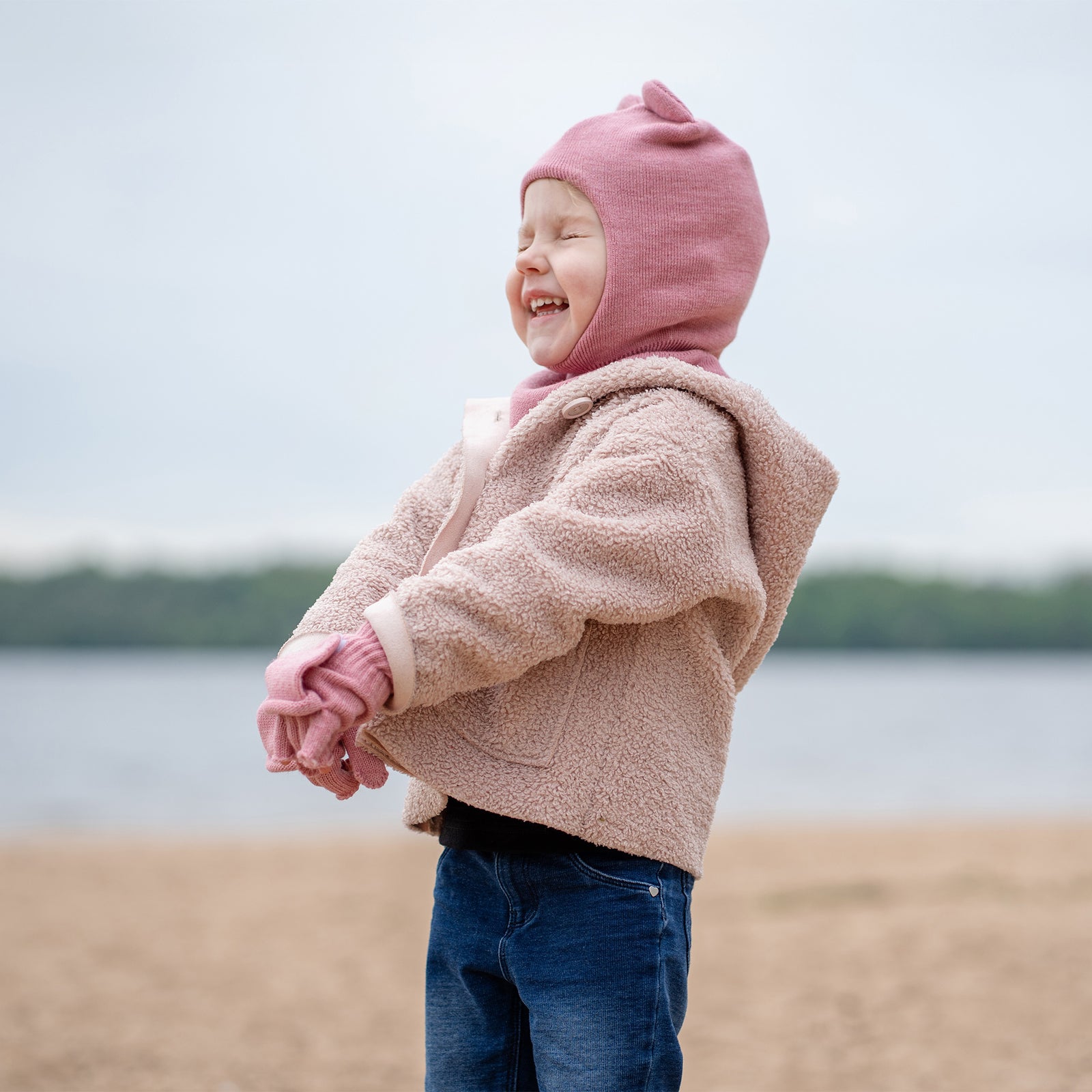 Un niño pequeño con ojos azules y una amplia sonrisa feliz, está sentado con las piernas cruzadas sobre una superficie blanca. Lleva una pasamontañas rosa con orejas, manoplas rosas a juego y una camisa y pantalones de manga larga de color claro.