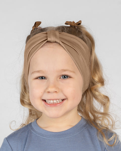 A close-up studio portrait of a young blonde girl smiling brightly at the camera against a white background. She is wearing a dusty blue short-sleeved t-shirt and a beige ribbon headband with a knot detail, with small brown hair bows visible behind it.