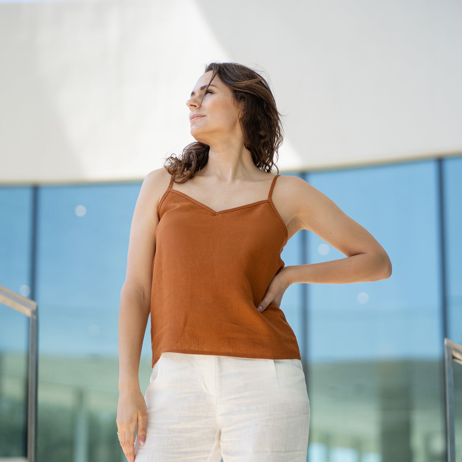 Una mujer de cabello castaño lleva una blusa de tirantes con escote en V color almendra y pantalones blancos, posando en el interior frente a grandes ventanales de cristal.