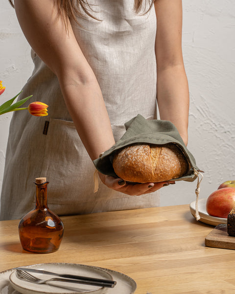 Una persona con un delantal de lino sostiene una barra de pan sobre una mesa de madera junto a otros alimentos.