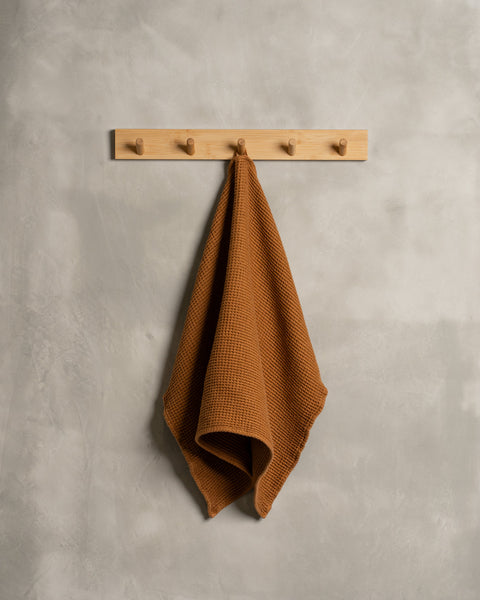 A gray wall with a wooden hanger. In the middle of it hangs a waffle hand washcloth in almond brown color.