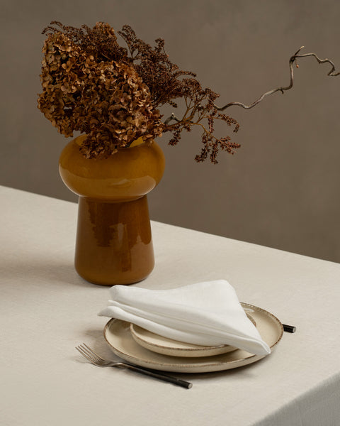 A tabletop covered with a linen tablecloth, with a vase and a single plate set on top of it. By the sides of the plates there are cutlery items, and there is a linen napkin in pure white color on top.