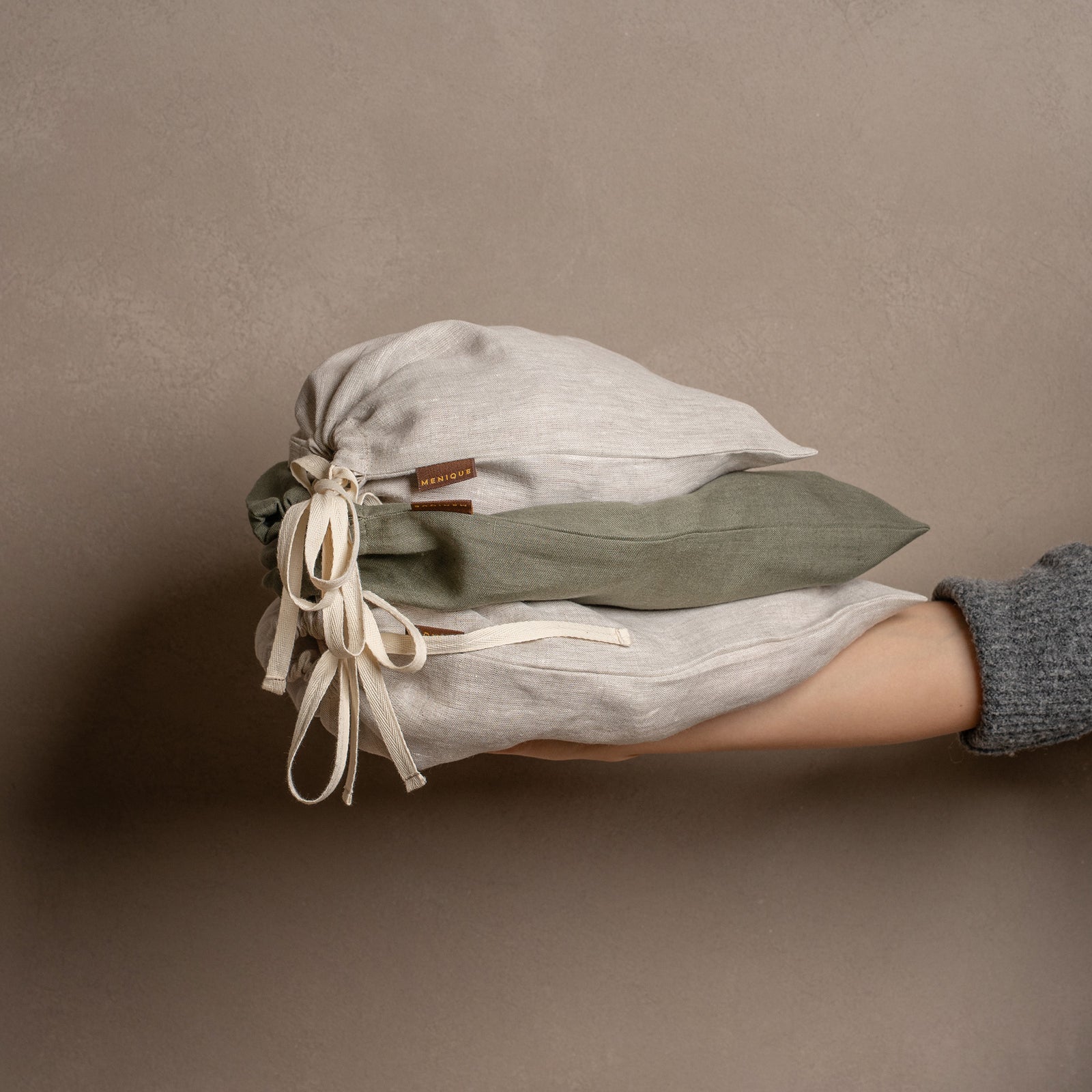 Cinco bolsas cerradas de lino con cordón, de diferentes tamaños y colores natural y verde piedra, están dispuestas sobre una alfombra de yute texturizada frente a una pared marrón cálida.