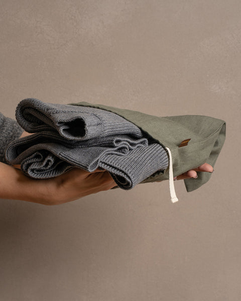 Las manos de una persona sostienen con delicadeza una prenda gris de punto doblada dentro de una bolsa de lino verde piedra con cordón, sobre un fondo marrón apagado.