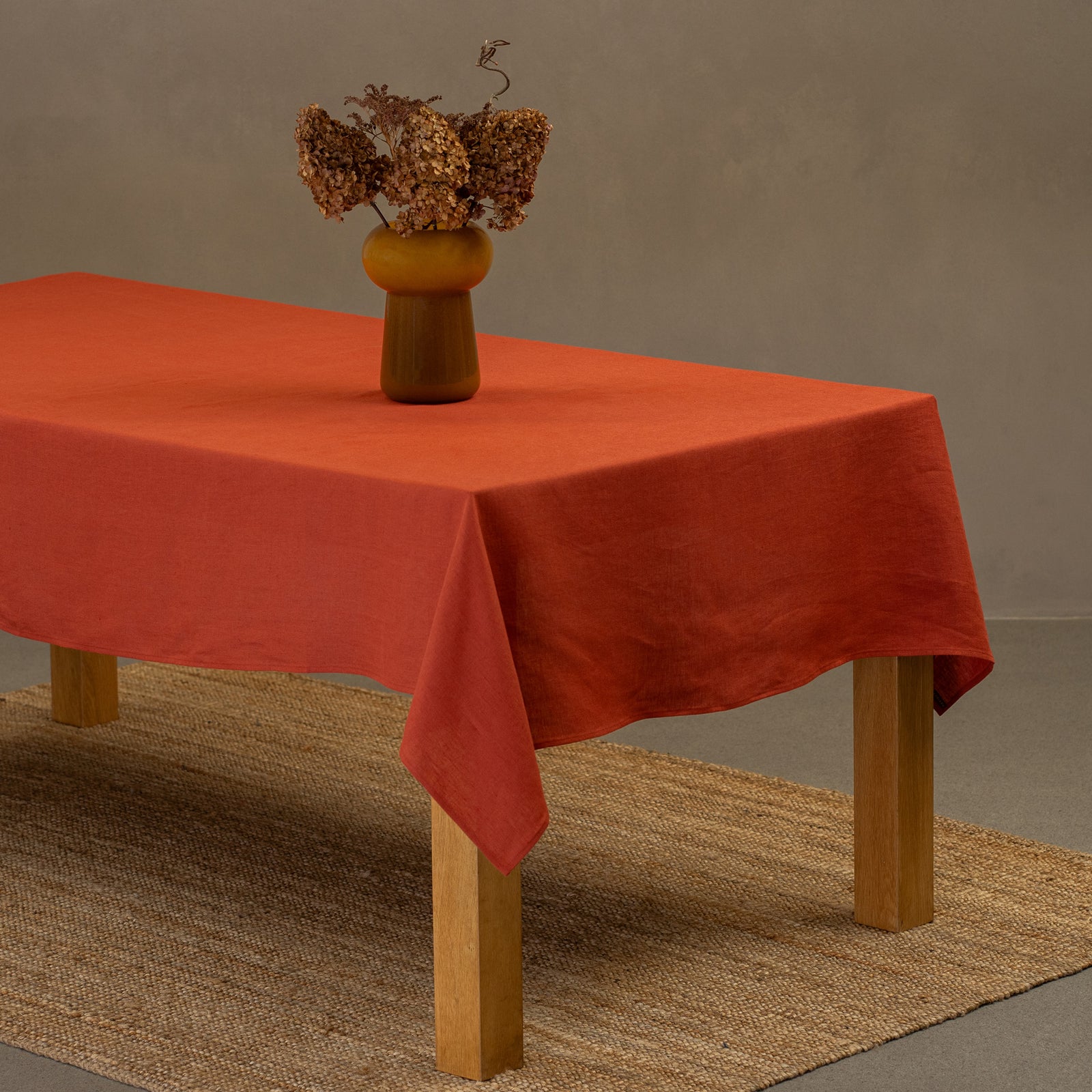 El mantel de lino verde piedra de menique sobre una mesa de madera. Encima del mantel hay un jarrón hecho de pajillas naturales y una taza de barro. Debajo de la mesa hay una alfombra.