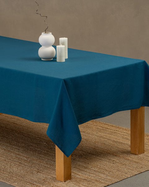 El mantel de lino menique en color azul cobalto, cayendo sobre una mesa de madera, con un jarrón blanco moderno encima y dos velas blancas de diferente altura a los lados.